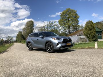 Een zilveren Toyota Highlander SUV staat geparkeerd op een landelijke weg met bomen, een kas en een blauwe lucht met verspreide wolken op de achtergrond.