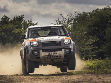 Een Bowler Defender Challenge-auto met een "DC11 BOW"-kentekenplaat rijdt op een bewolkte dag met hoge snelheid over een stoffige onverharde weg, omringd door bomen.