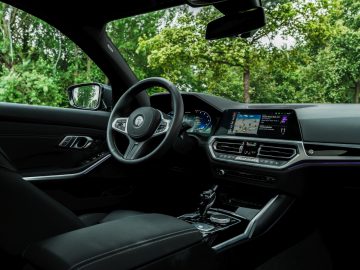 Binnenaanzicht van een moderne BMW Alpina met de bestuurdersstoel, het stuur, het dashboard met digitaal display en groene bomen zichtbaar door de ramen.
