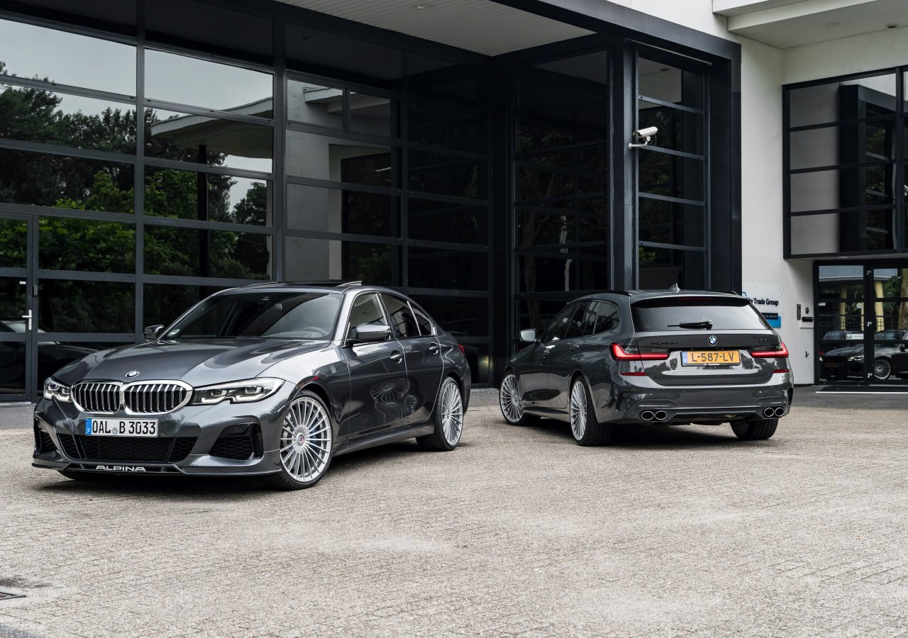 Twee grijze BMW Alpina-auto's staan geparkeerd buiten een modern gebouw met grote glazen ramen. De auto aan de linkerkant is een sedan en de auto aan de rechterkant is een stationwagen.