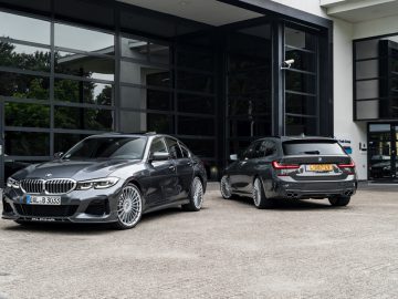 Twee grijze BMW Alpina-auto's staan geparkeerd buiten een modern gebouw met grote glazen ramen. De auto aan de linkerkant is een sedan en de auto aan de rechterkant is een stationwagen.