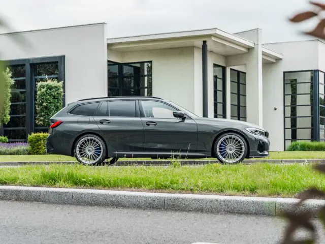 Voor een modern wit gebouw met grote ramen en een groen gazon staat een strakke BMW Alpina stationwagen geparkeerd.