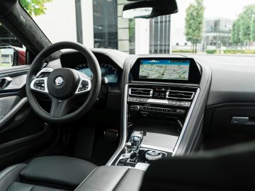 Binnenaanzicht van een moderne BMW Alpina met een stuur, digitaal display en navigatiesysteem, met gebouwen en groen zichtbaar buiten de ramen.