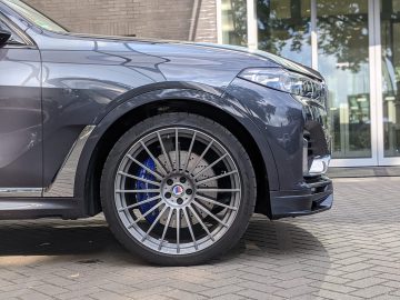 Close-up van het voorwiel en spatbord van een donkerblauwe BMW Alpina luxe SUV, geparkeerd op een verhard oppervlak voor een gebouw met glazen deuren. De auto is voorzien van grote, meerspaaks lichtmetalen velgen.