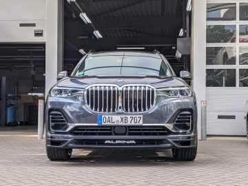Vooraanzicht van een metaalgrijze BMW Alpina SUV geparkeerd voor een garage, waarbij de kenmerkende grille en koplampen prominent zichtbaar zijn.