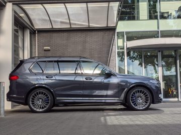 Een grijze BMW Alpina SUV staat geparkeerd op een verhard oppervlak voor een gebouw met grote ramen en een overstek.