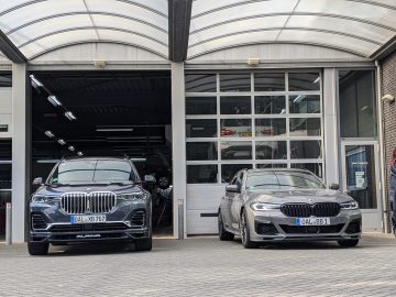 Twee BMW Alpina-auto's, een zwarte en een grijze, geparkeerd voor een autoservicegarage met grote, open deuren en doorzichtige ramen.