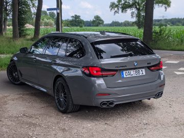 Op een grindweg bij een T-splitsing staat een strakke grijze BMW Alpina B5 geparkeerd, omgeven door bomen en een open veld. De auto, voorzien van het Duitse kenteken "OAL-BB 1", valt op in zijn schilderachtige landelijke omgeving.