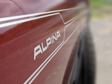 Close-up van een kastanjebruine BMW Alpina-auto met witte 'Alpina'-tekst en streepdetails op de zijkant.