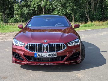 Vooraanzicht van een strakke rode BMW Alpina, geparkeerd op een verhard terrein, perfect omlijst door weelderige groene bomen op de achtergrond.