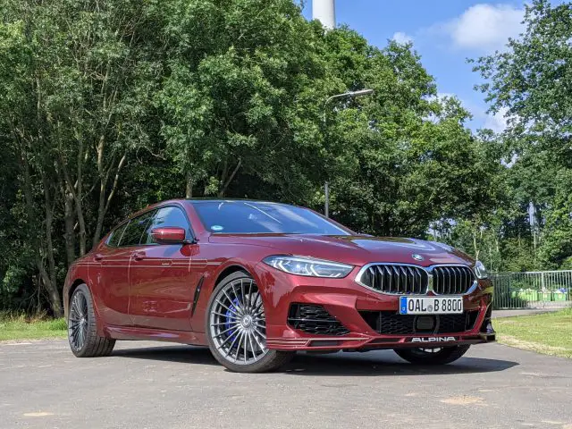 Een rode BMW Alpina B8 sedan staat geparkeerd op een verhard oppervlak, met bomen en een straatlantaarn op de achtergrond, wat het strakke design en de elegantie van de BMW Alpina-serie benadrukt.