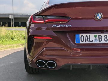 Achteraanzicht van een kastanjebruine BMW Alpina-auto met dubbele uitlaten, een Europees kenteken met de tekst "OAL B 8" en een serene watermassa en een pittoreske brug op de achtergrond.