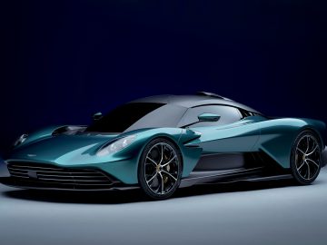 Tegen een donkere achtergrond staat een slanke, blauwgroenkleurige Aston Martin Valhalla-sportwagen met een laag, aerodynamisch ontwerp en zwarte accenten.