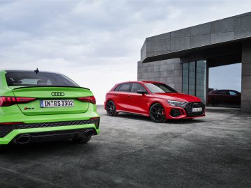 Twee Audi RS 3 auto's in een buitenstedelijke omgeving. De ene auto is groen en kijkt van de camera af, terwijl de andere rood is en naast een modern gebouw geparkeerd staat.