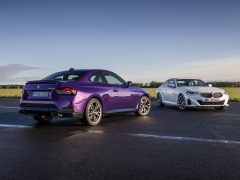 Twee auto's geparkeerd op een verhard oppervlak. De ene is een paarse sportwagen die wegkijkt, en de andere is een witte BMW 2 Serie Coupé die naar de kijker kijkt. Open veldachtergrond onder een heldere hemel.