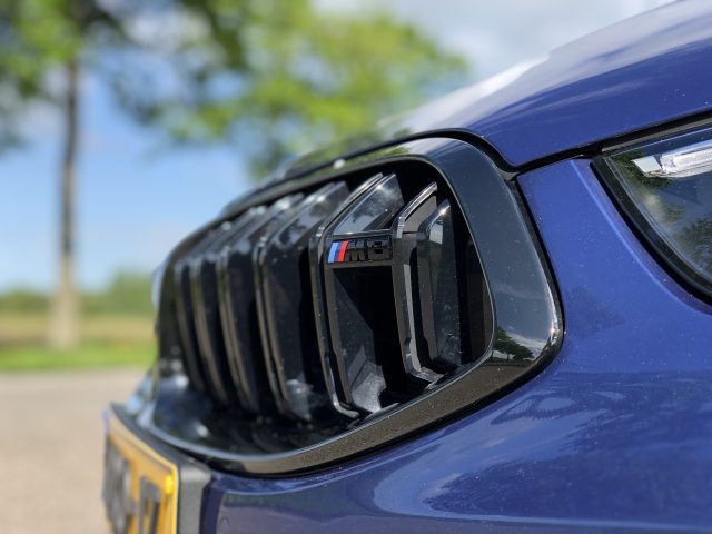 Close-up van de grille van een blauwe BMW M8 Gran Coupé Competition met het M-logo zichtbaar, buiten geparkeerd op een zonnige dag. Bomen en lucht zijn enigszins wazig op de achtergrond.