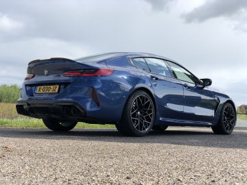 Een blauwe BMW M8 Gran Coupé Competition met Nederlands kenteken staat geparkeerd op een verharde ondergrond, achteraanzicht onder een bewolkte hemel.