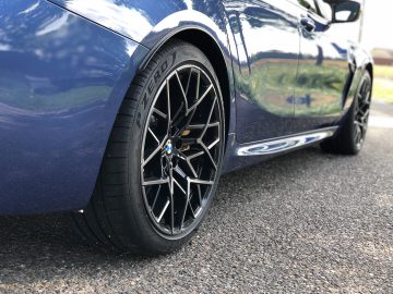 Close-up van een blauwe BMW M8 Gran Coupé Competition, geparkeerd op een weg, met de nadruk op het linkervoorwiel met een zwarte lichtmetalen velg en Pirelli P Zero-band. Reflecties van de omgeving zijn te zien in het gepolijste oppervlak van de auto.