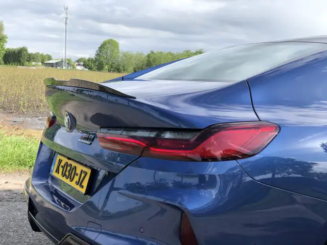 Een close-up van de achterkant van een blauwe BMW M8 Gran Coupé Competition met een zichtbaar geel kenteken en opvallend achterlichtontwerp, geparkeerd nabij een veld met bomen en een communicatietoren op de achtergrond.