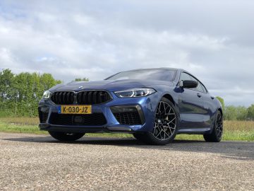 Een blauwe BMW M8 Gran Coupé Competition geparkeerd op een weg met een gedeeltelijk bewolkte lucht en groen gebladerte op de achtergrond.