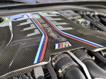 Close-up van een krachtige motor met 'BMW M Power'-logo en M-Series-logo, met koolstofvezeldetails en blauwe en rode strepen, onder de motorkap van de BMW M8 Gran Coupé Competition.