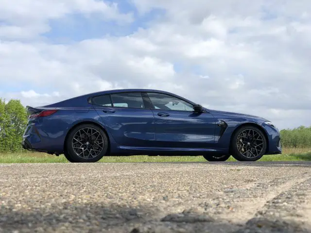 Een zijaanzicht van een blauwe BMW M8 Gran Coupé Competition geparkeerd op een verhard oppervlak met gras en bomen op de achtergrond onder een gedeeltelijk bewolkte hemel.