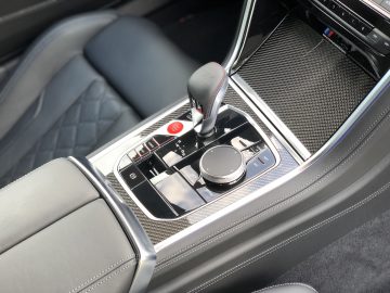 Close-up van de middenconsole van een BMW M8 Gran Coupé Competition met een versnellingspook, verschillende bedieningsknoppen, een draaiknop en sieraccenten van koolstofvezel. De stoelen zijn voorzien van gewatteerde lederen bekleding.