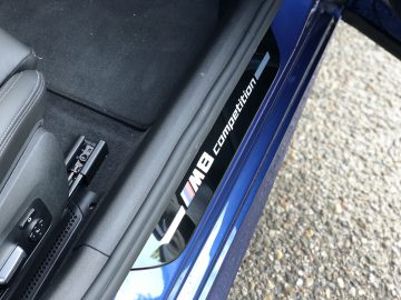 Close-up van de dorpel van een auto met het 'M8 competition'-logo, naast een deel van een gestructureerd grondoppervlak, wat de luxueuze details van de BMW M8 Gran Coupé Competition benadrukt.