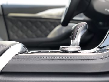 Close-up van de middenconsole van een moderne auto met een versnellingspook met contrasterende stiksels en een draaiknop, die de luxueuze details van de BMW M8 Gran Coupé Competition vastlegt. Op de achtergrond is een deel van de zitting met gewatteerde bekleding te zien.