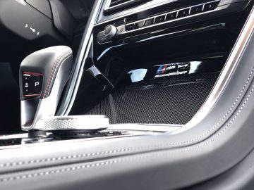 Close-up van een luxueus auto-interieur met een versnellingspook, een bedieningsknop en een badge met "TFSI competition" op een glanzend zwart paneel, dat doet denken aan het premiumgevoel van de BMW M8 Gran Coupé Competition.