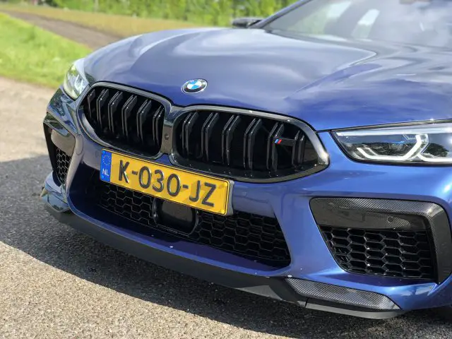 Close-up van de voorkant van een blauwe BMW M8 Gran Coupé Competition met een geel kenteken met de tekst "K-030-JZ", geparkeerd op een verhard oppervlak met groen op de achtergrond.