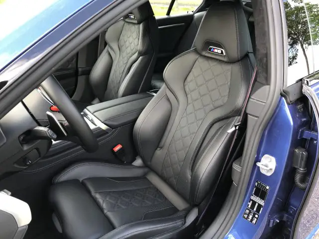 Interieur van een BMW M8 Gran Coupé Competition met zwartleren stoelen met een ruitvormig stikselpatroon, en de bestuurdersstoel met een M-badge. De autodeur staat open, waardoor delen van de buitenkant blauw geverfd zijn.