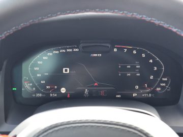 Close-up van een digitaal autodashboard in een BMW M8 Gran Coupé Competition met een snelheid van 0 km/u. Het display toont verschillende indicatoren, waaronder afstand, brandstofmeter, temperatuur en waarschuwingslampjes.