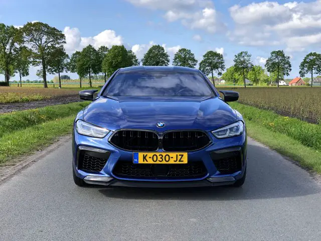 Een blauwe BMW M8 Gran Coupé Competition met Nederlands kenteken K-030-JZ staat geparkeerd op een landelijke weg, omgeven door groene velden en bomen, onder een gedeeltelijk bewolkte hemel.