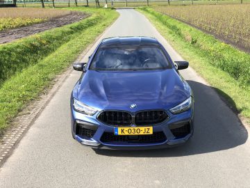 Op een smal landweggetje, omzoomd door groen gras en landbouwgrond, staat een blauwe BMW M8 Gran Coupé Competition met Nederlands kenteken "K-030-JZ" geparkeerd.