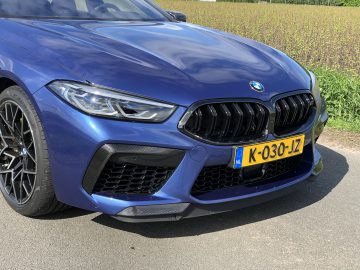 Close-up van de voorkant van een blauwe BMW M8 Gran Coupé Competition met zichtbaar Nederlands kenteken met de tekst "K-030-JZ", geparkeerd op een weg naast een veld.