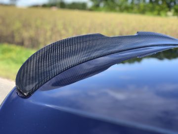 Close-up van een koolstofvezelspoiler op een BMW M8 Gran Coupé Competition voor een wazig grasveld en bomen.