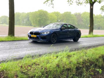 Een blauwe BMW M8 Gran Coupé Competition rijdt op een landelijke weg omgeven door bomen en gras.