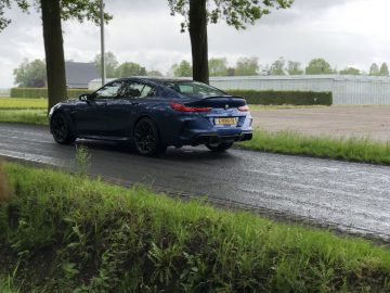 Een blauwe BMW M8 Gran Coupé Competition rijdt op een smalle, natte weg omzoomd met bomen. De kentekenplaat is zichtbaar en er zijn grasvelden en een omheind gebied op de achtergrond onder een bewolkte hemel.