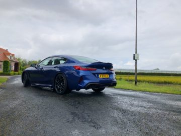 Een blauwe BMW M8 Gran Coupé Competition met kenteken "K-430-JZ" staat geparkeerd op een natte plattelandsweg naast een veld en een huis met een rood dak onder een bewolkte hemel.