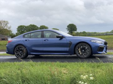 Een blauwe BMW M8 Gran Coupé Competition sedan staat geparkeerd aan de kant van een weg met een met gras begroeide voorgrond en bomen op de achtergrond onder een bewolkte hemel.