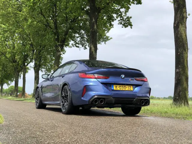 Op een met bomen omzoomde weg staat een blauwe BMW M8 Gran Coupé Competition met kenteken K-030-JZ geparkeerd. De foto is genomen vanaf de achterkant van het voertuig.