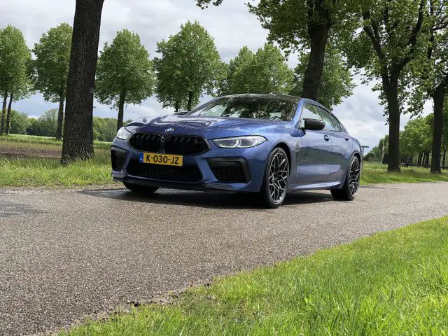 Een blauwe BMW M8 Gran Coupé Competition geparkeerd op een smalle, met bomen omzoomde weg op een bewolkte dag, met groene velden en bomen op de achtergrond.