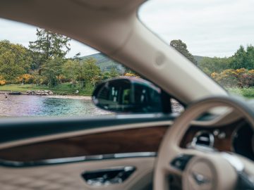 Uitzicht vanuit een auto, met het strakke ontwerp van de Lotus Emira op de voorgrond, met een landschap aan het meer met bomen en een persoon die bij het water staat.