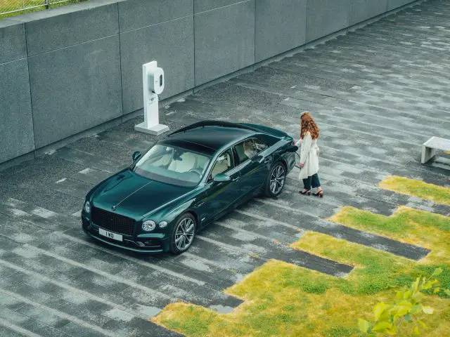 Een persoon met lang haar staat naast een donkergroene Lotus Emira, geparkeerd voor een laadstation voor elektrische voertuigen op een moderne, verharde buitenruimte.