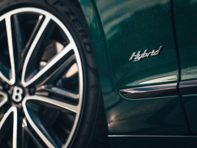 Close-up van een Lotus Emira met een groene afwerking, met een wiel versierd met de letter "B" en een "Hybrid"-badge op de zijkant.