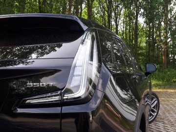 Close-up van een zwarte Cadillac XT4 met een '350T'-badge op de achterkant, geparkeerd op een verhard terrein nabij een bos. De afbeelding is genomen vanaf de linkerachterzijde en laat het ontwerp van het achterlicht en de reflecterende omgeving zien.