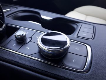 Close-up van de console van een Cadillac XT4 met een grote bedieningsknop, een aangrenzende kleinere knop en verschillende knoppen voor verschillende functies. De console is voorzien van bekerhouders en heeft een zwart lederen afwerking.