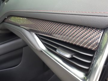 Close-up van een Cadillac XT4-interieur met een dashboardafwerking met koolstofvezelpatroon, een zilveren ventilatieopening en een zwart lederen oppervlak met rode stiksels.