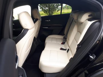 De achterbank van de Cadillac XT4, versierd met beige lederen bekleding en zichtbare veiligheidsgordels, biedt voldoende beenruimte en door de ramen zicht op het groen buiten.